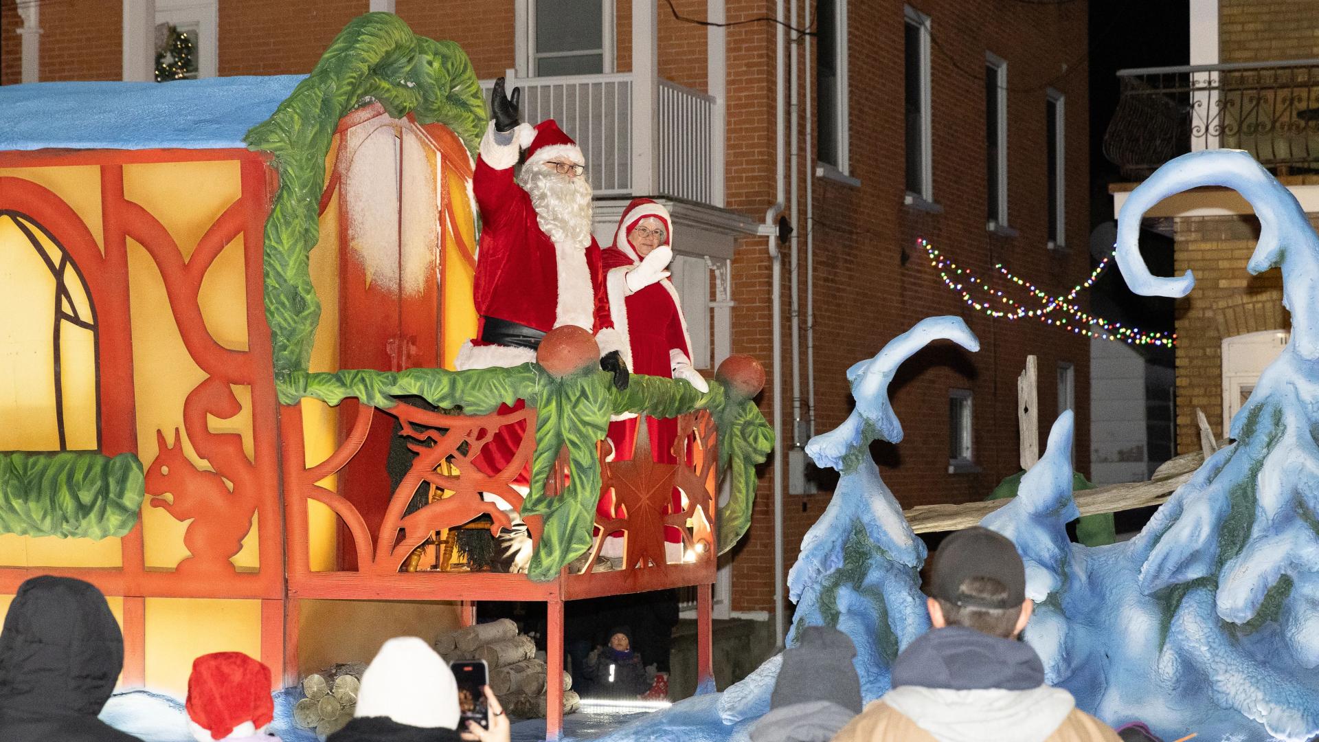 Santa Claus and Mrs. Claus waving from a brightly lit Christmas parade float decorated with winter scenery and holiday lights.